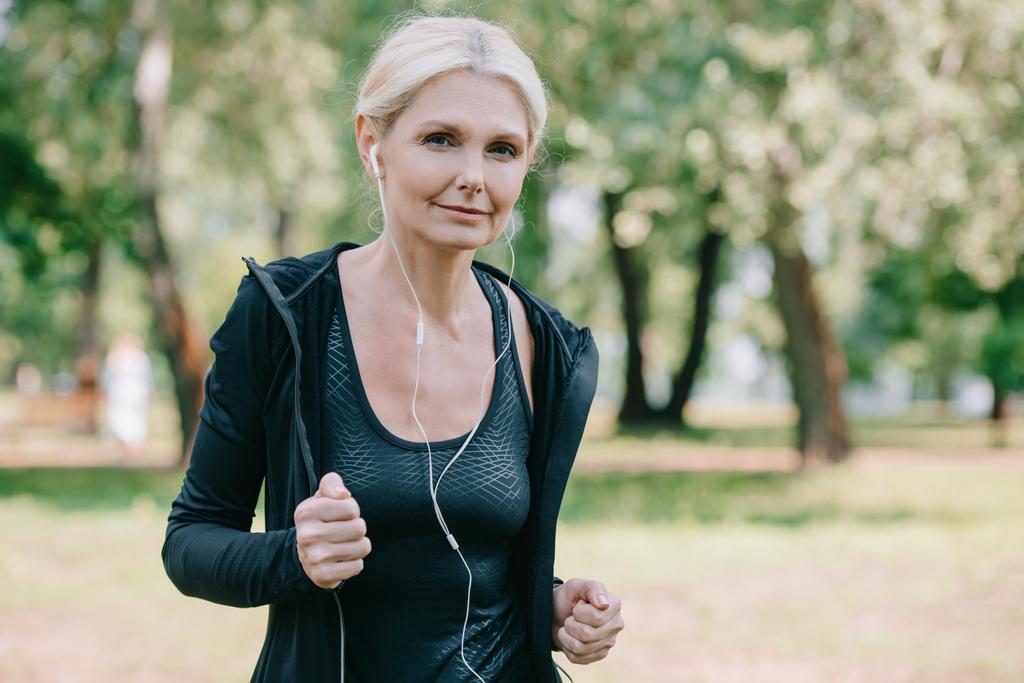 beautiful mature sportswoman smiling at camera while running in park and listening music in earphones - Photo, Image