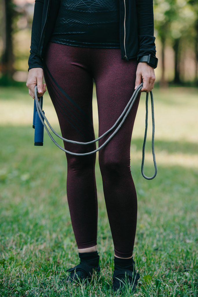 обрізаний вид зрілої спортсменки, що стоїть в парку і тримає стрибкову мотузку
 - Фото, зображення