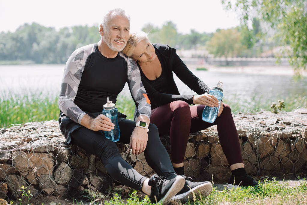 Lächelnder älterer Sportler und Sportlerin, die auf Brüstung im Park sitzen und Sportflaschen in der Hand halten - Foto, Bild