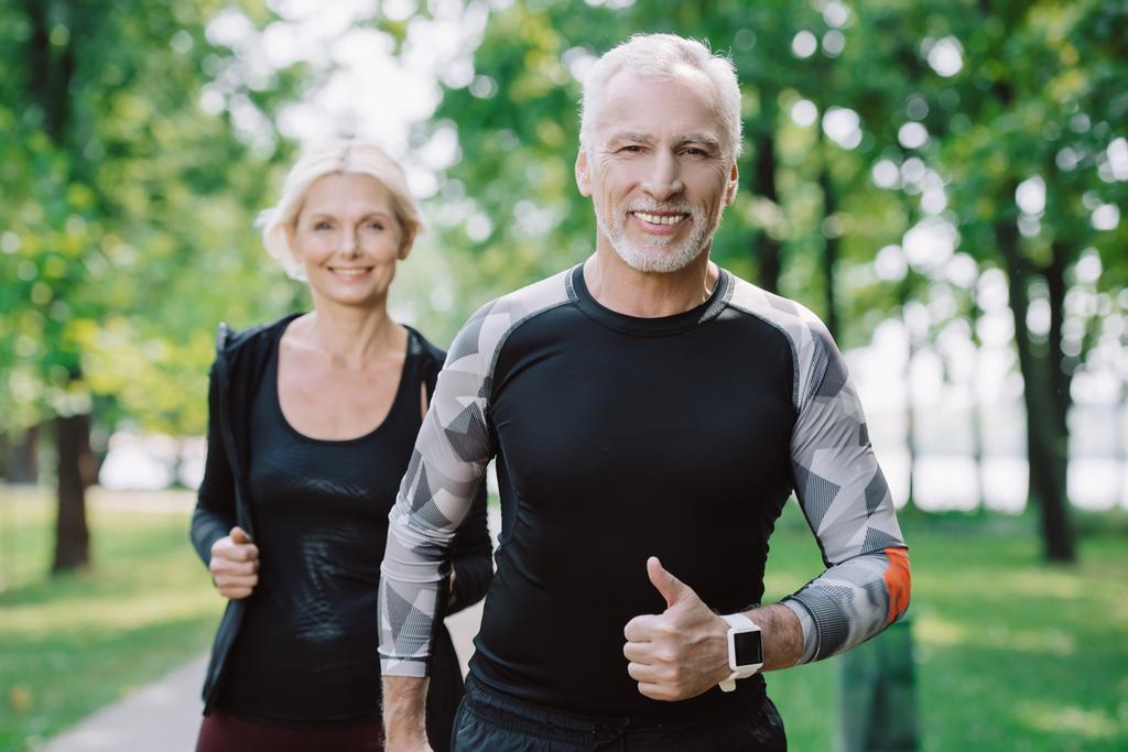 cheerful mature sportsman showing thumb up while jogging in park together with smiling sportswoman - Photo, Image