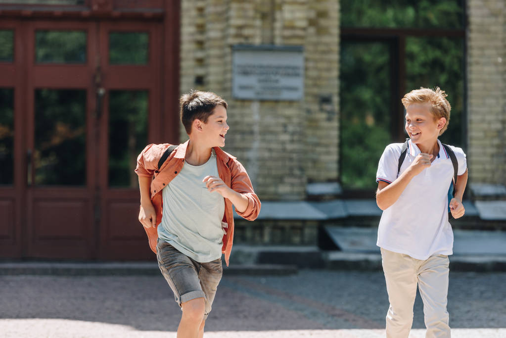 kaksi söpöä koulupoikaa juoksemassa koulun pihalla hymyillen ja katsoen toisiaan
  - Valokuva, kuva