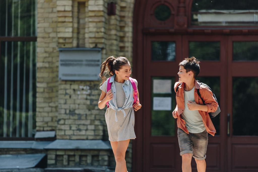 веселый школьник и школьница улыбаются и смотрят друг на друга во время бега по школьному двору
 - Фото, изображение
