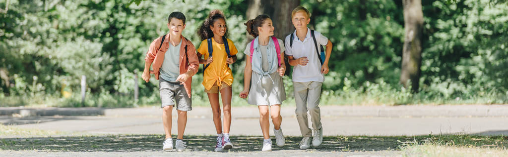 panoramische foto van vier gelukkige multiculturele schoolkinderen die in Sunnypark rennen - Foto, afbeelding
