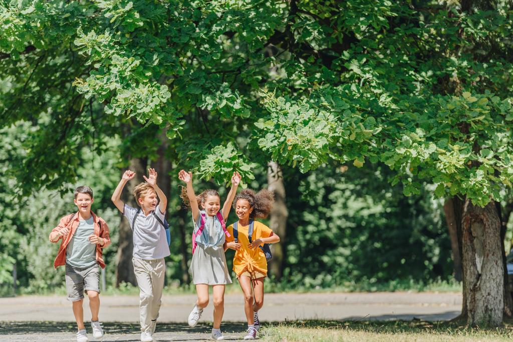 four happy multicultural schoolkids gesturing with raised hands while running in park - Photo, Image