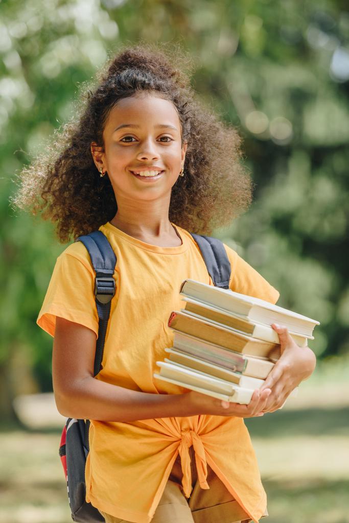 Gelukkig African American schoolmeisje houdt boeken en glimlachend op camera - Foto, afbeelding