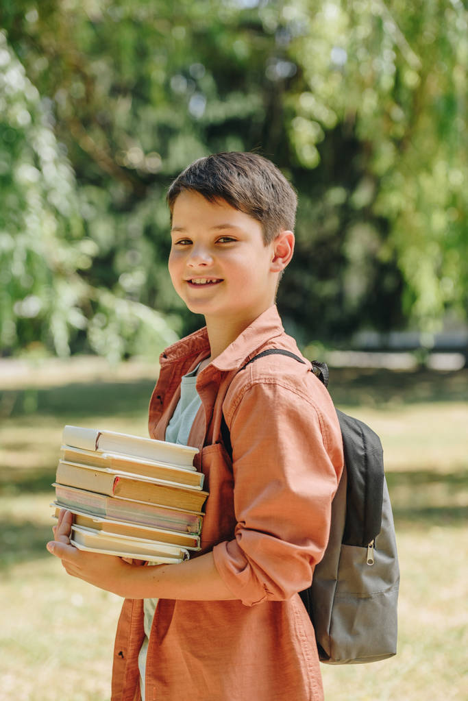 happy schoolboy smiling at camera while holding books in park - Photo, Image