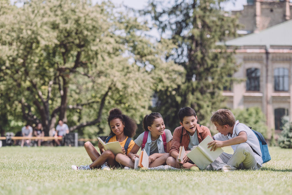 чотири веселі мультикультурні школярі сидять на газоні і читають книги
 - Фото, зображення