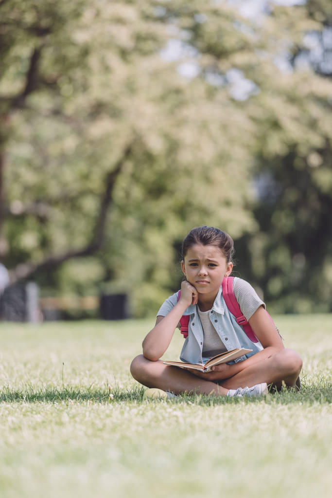 cute, bored schoolgirl holding book while sitting on lawn in park - Photo, Image
