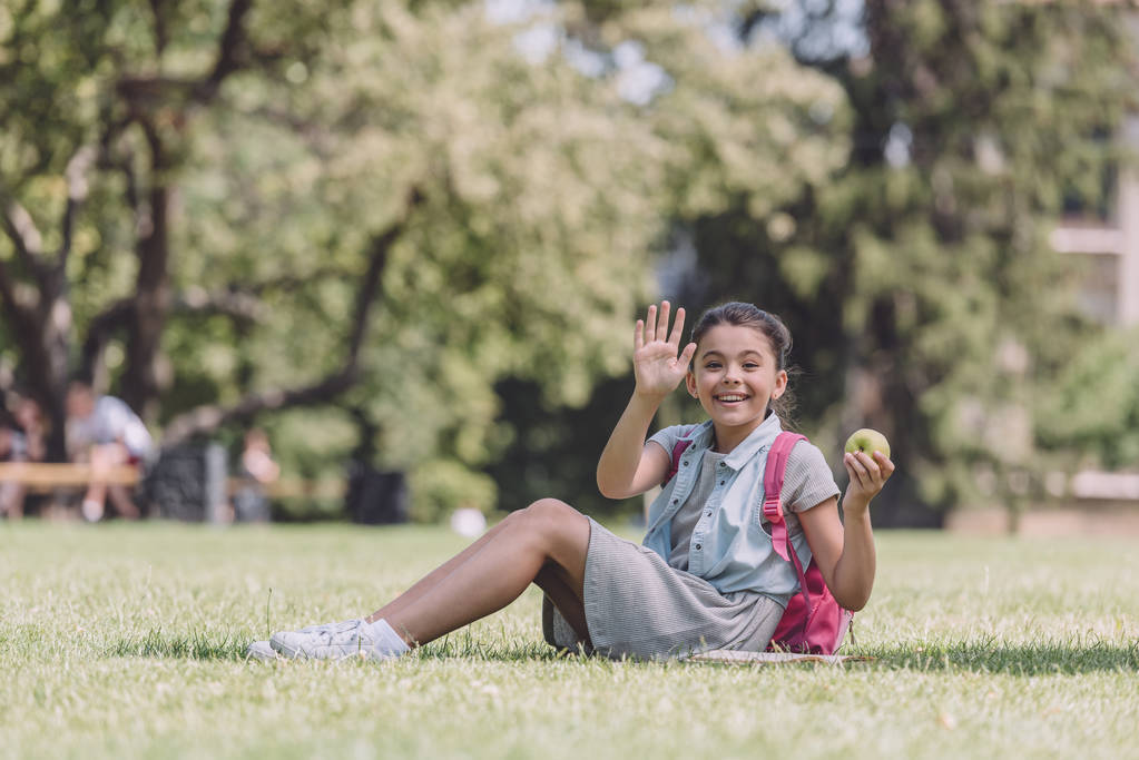 fröhliche Schülerin winkt mit der Hand, während sie auf dem Rasen im Park sitzt und in die Kamera schaut - Foto, Bild