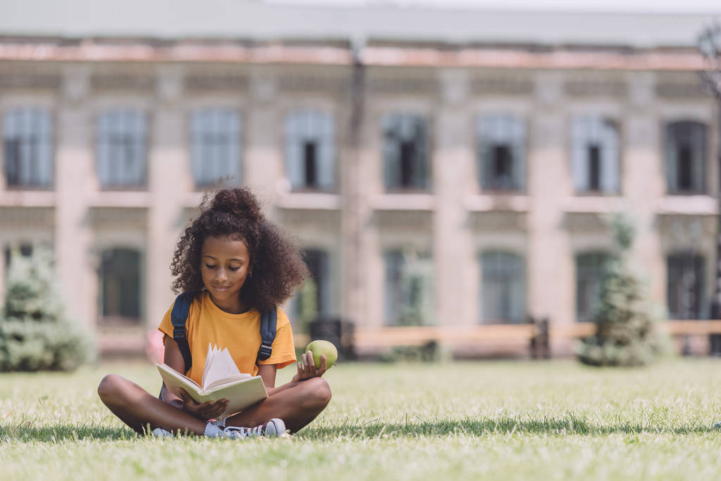 sonriente afroamericana colegiala sosteniendo manzana y leer libro mientras está sentado en el césped
 - Foto, imagen