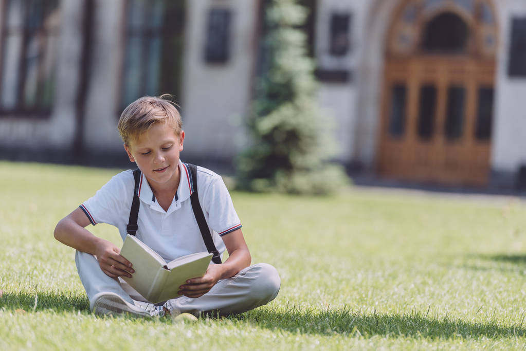 adorable écolier assis sur la pelouse près de l'école et livre de lecture
 - Photo, image