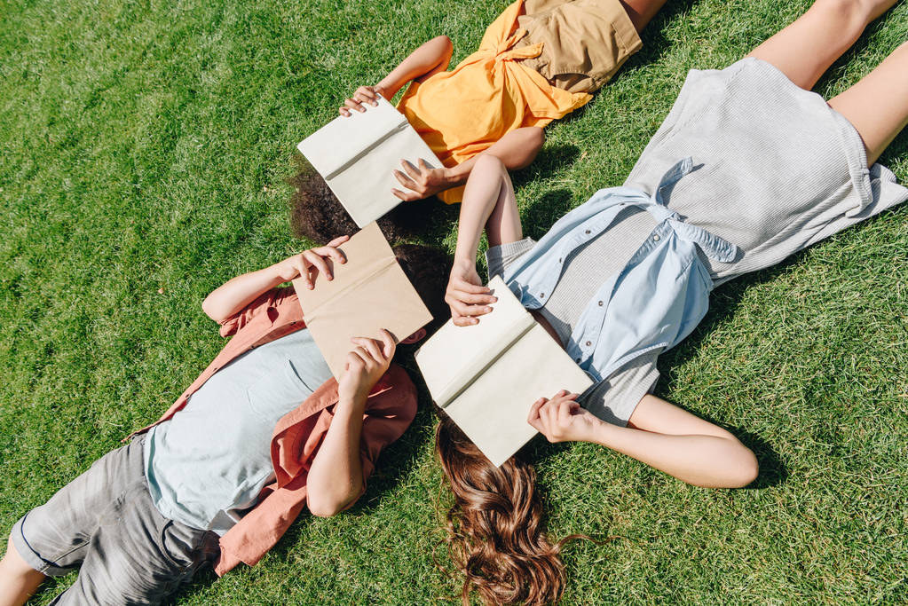 vista superior de tres escolares multiculturales tumbados en el césped y cubriendo caras con libros
 - Foto, Imagen