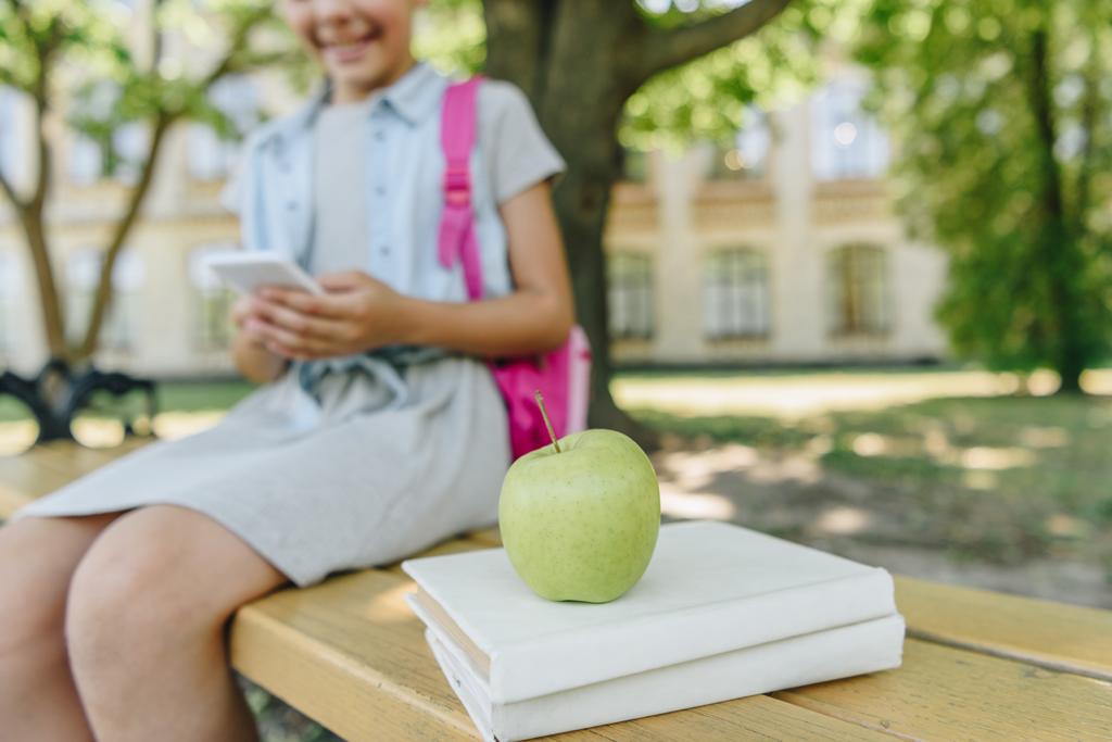 przycięte widok uśmiechniętą uczennicę za pomocą smartfona siedząc na ławce w pobliżu książek i jabłek - Zdjęcie, obraz