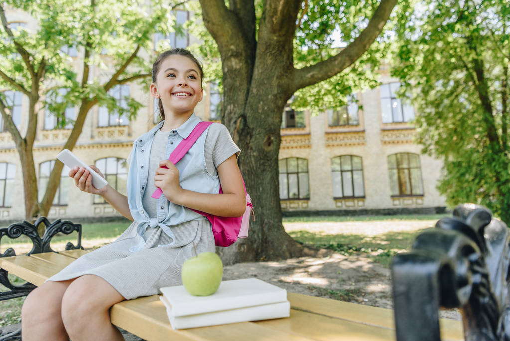 χαρούμενο μαθήτρια κρατώντας smartphone και κοιτάζοντας μακριά, ενώ κάθονται στο παγκάκι στο πάρκο - Φωτογραφία, εικόνα