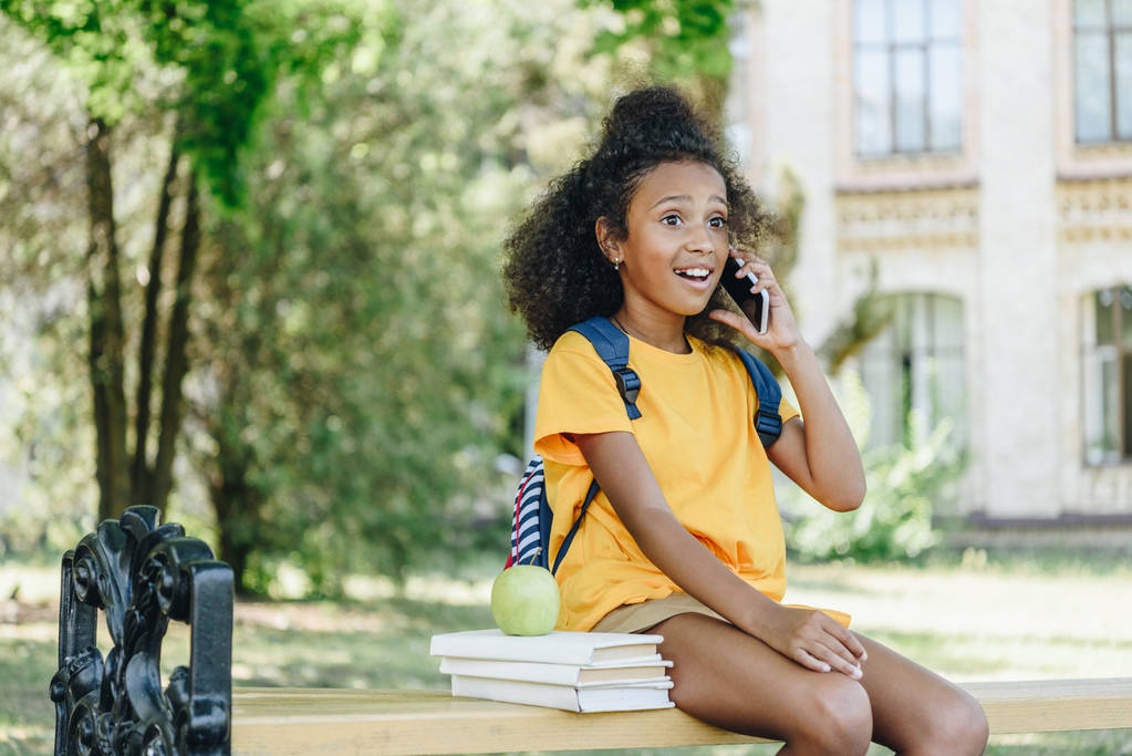 sorprendió a la chica afroamericana hablando en un teléfono inteligente mientras estaba sentada en el banco cerca de libros y manzana
 - Foto, imagen