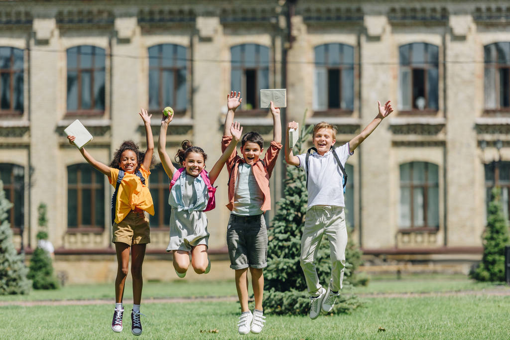 quattro scolari multiculturali felici che saltano mentre tengono libri sul prato nel parco
 - Foto, immagini