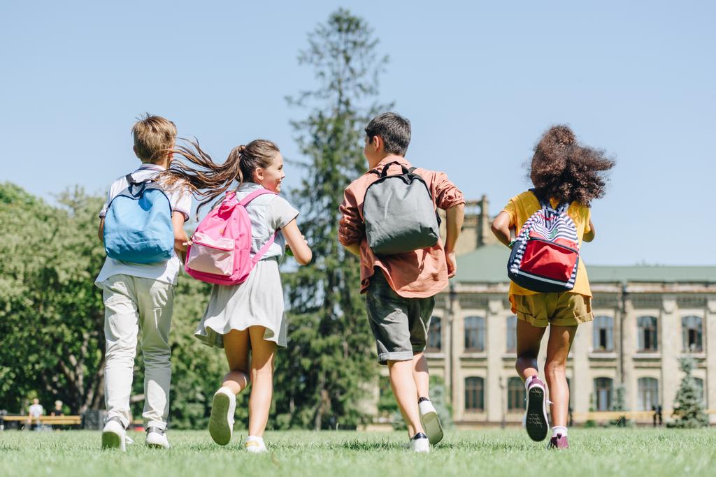 vista trasera de cuatro escolares multiétnicos con mochilas corriendo en el césped en el parque
 - Foto, imagen