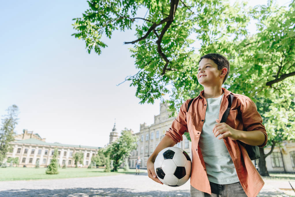 милый школьник с футбольным мячом прогулка в парке с голубым небом и зеленым деревом на заднем плане
 - Фото, изображение
