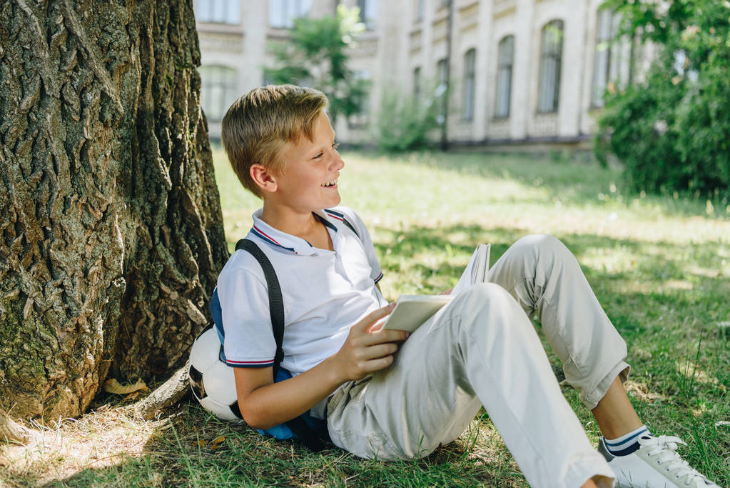 adorabile sorridente scolaro seduto sul prato sotto l'albero e tenendo libro
 - Foto, immagini
