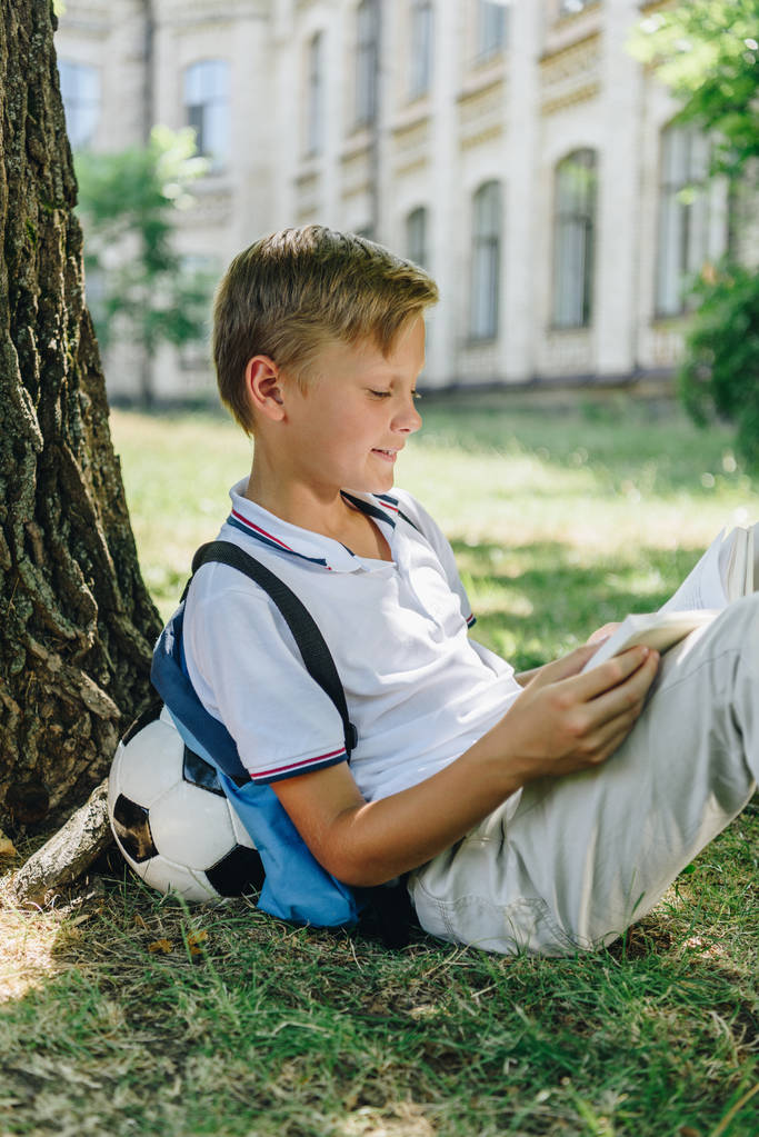 hymyilevä koulupoika lukemassa kirjaa istuessaan nurmikolla puun alla
 - Valokuva, kuva