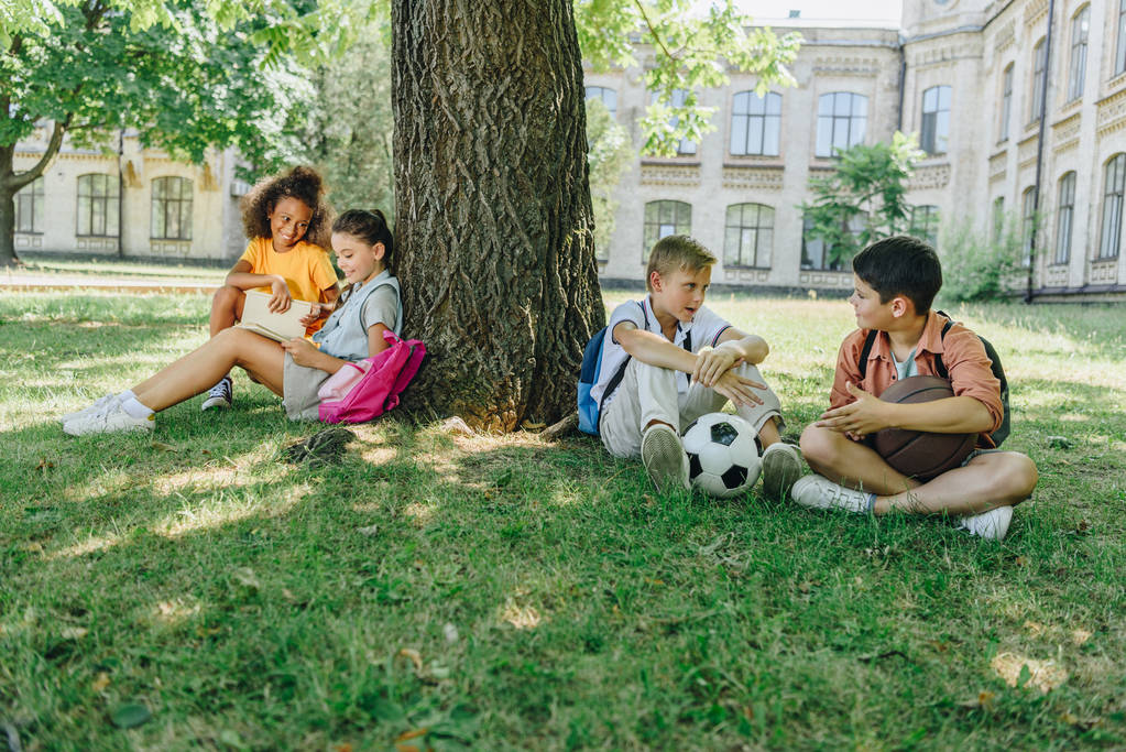 четыре милые мультикультурные школьники сидят на газоне и дереве, говорят и читают книги
 - Фото, изображение