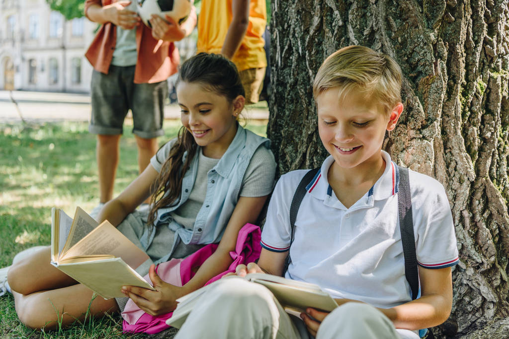 entzückende Schulkinder sitzen auf dem Rasen und lesen Bücher bei multikulturellen Freunden - Foto, Bild