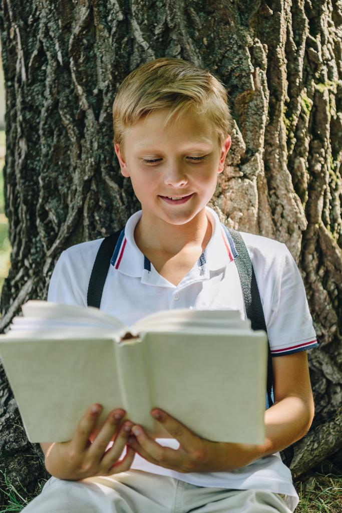 adorable sonriente colegial sentado cerca de árbol y libro de lectura
 - Foto, imagen