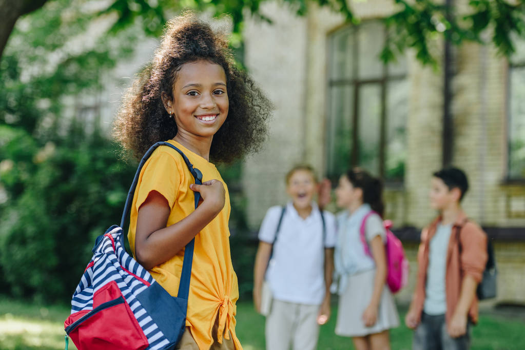 selective focus of cheerful african american schoolgirl with backpack smiling at camera near multiethnic friends - Photo, Image