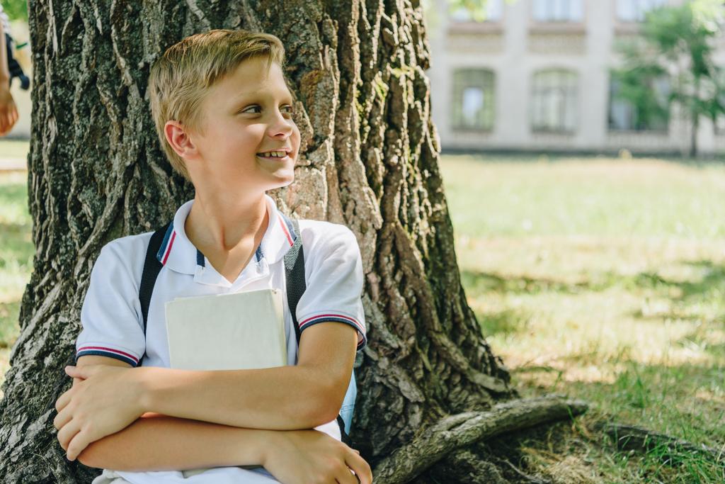 schattige school jongen zittend op gazon onder de boom, glimlachend en op zoek weg - Foto, afbeelding