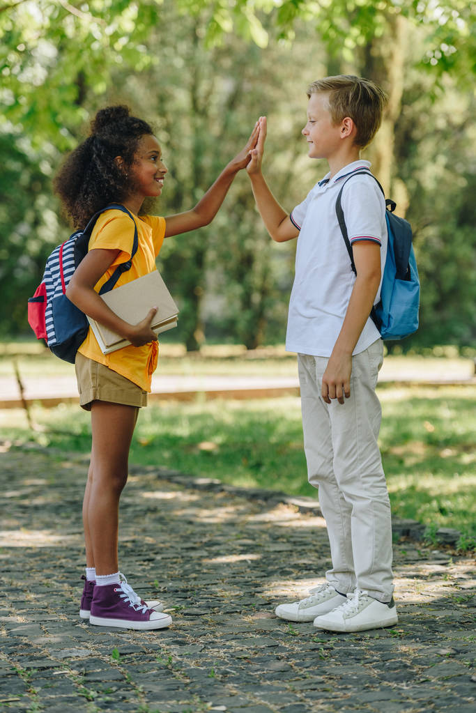 two smiling multicultural schoolkids giving hive five while standing in park - Photo, Image