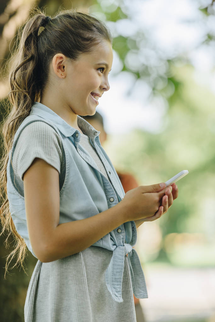 симпатичная улыбчивая школьница улыбается, отводя взгляд от смартфона в парке
 - Фото, изображение