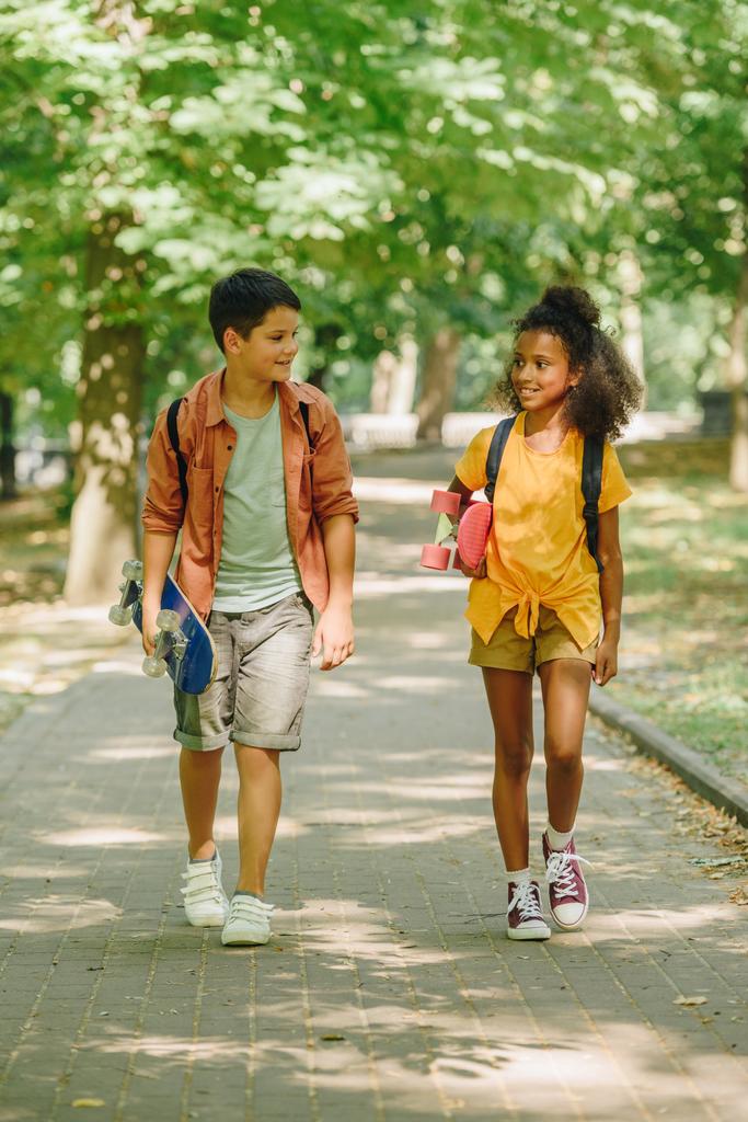 δύο πολυπολιτισμικούς μαθητές που περπατούν στο πάρκο ενώ κρατούν σκέιτμπορντ - Φωτογραφία, εικόνα