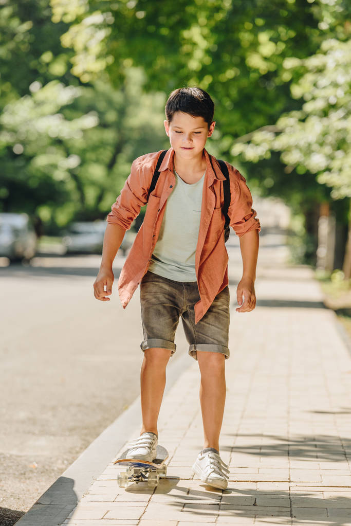cute schoolboy with backpack riding skateboard on sunny street  - Photo, Image