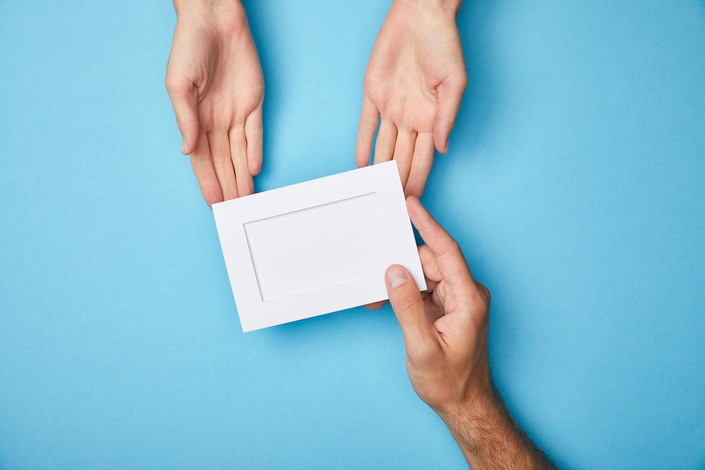 oříznutý pohled muže, který dává bílou prázdnou kartu do rámečku ženě na modrém pozadí - Fotografie, Obrázek