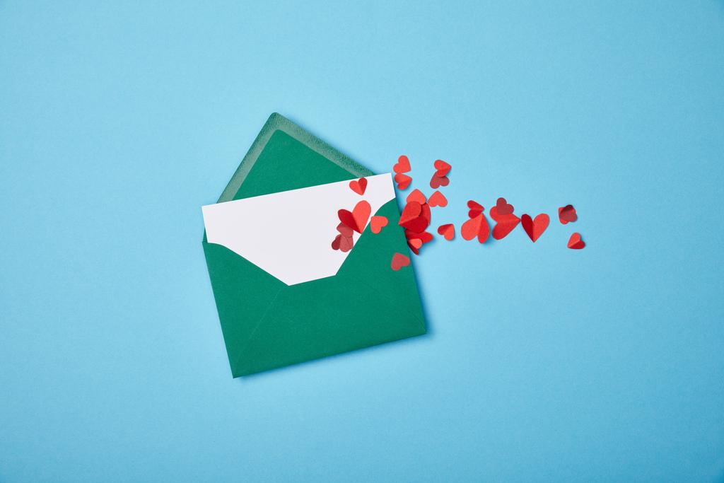 grüner Umschlag mit leerer weißer Karte und roten Papierherzen auf blauem Hintergrund - Foto, Bild