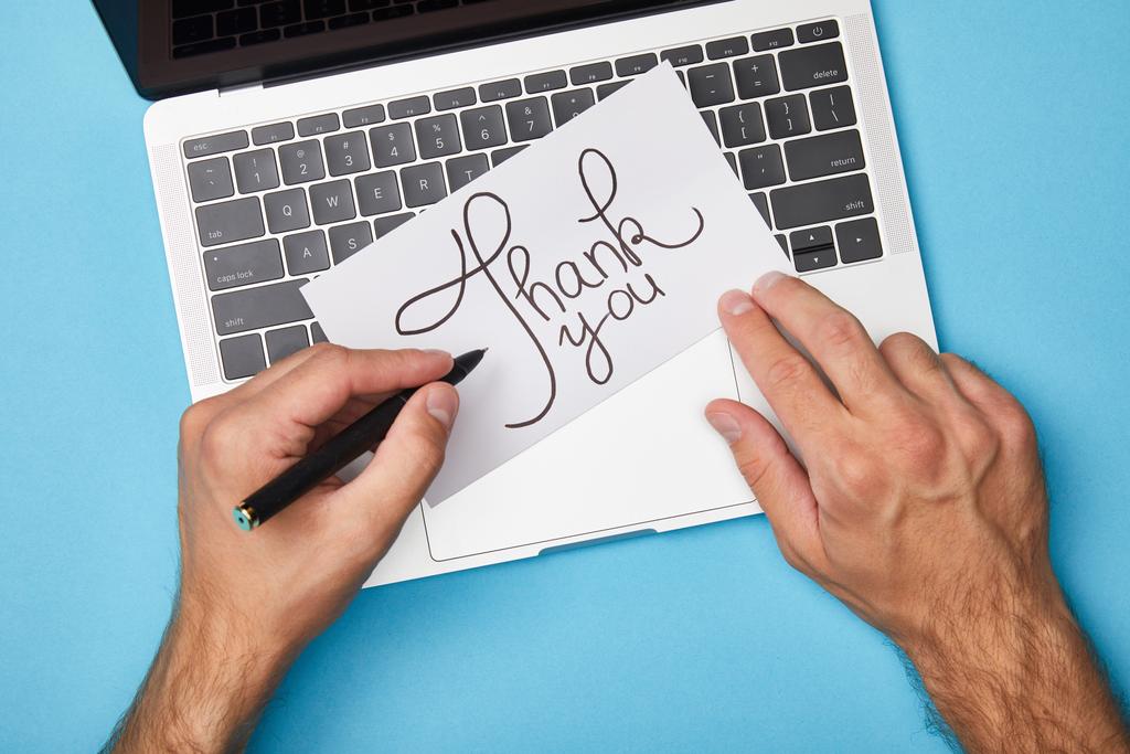 vista recortada del hombre escribiendo en la tarjeta con letras de agradecimiento cerca del ordenador portátil sobre fondo azul
 - Foto, Imagen