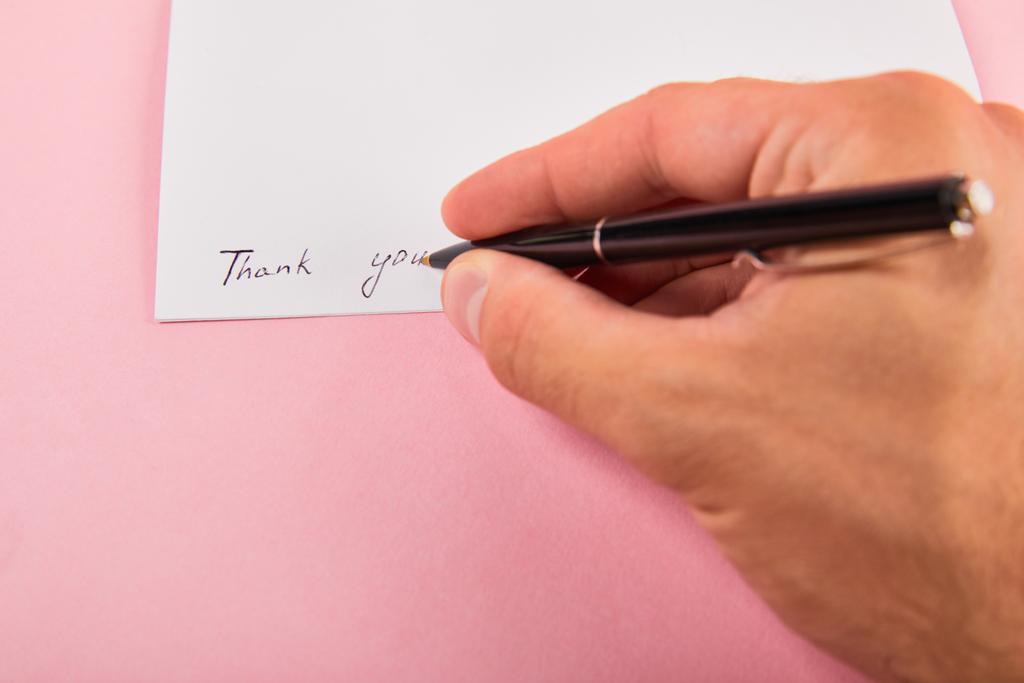 Ausgeschnittene Ansicht eines Mannes, der mit Stift Dankeschön-Schriftzug auf weißer Karte auf rosa Hintergrund schreibt - Foto, Bild