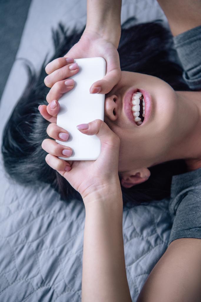 κατάθλιψη γυναίκα στο κρεβάτι καλύπτοντας το πρόσωπο με smartphone στο υπνοδωμάτιο - Φωτογραφία, εικόνα