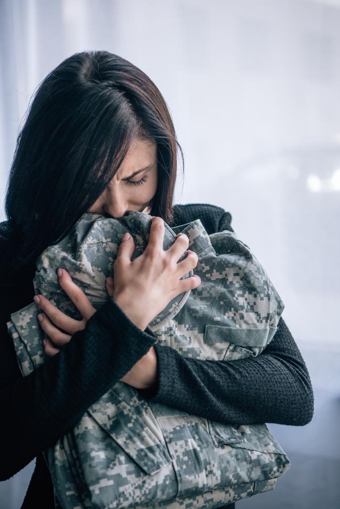 κατάθλιψη γυναίκα κλαίει και κρατώντας στρατιωτικά ρούχα στο σπίτι - Φωτογραφία, εικόνα
