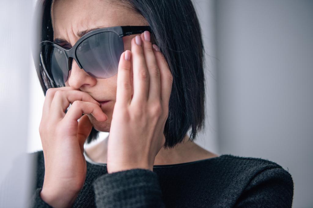 Kobieta z depresją w okulary przeciwsłoneczne obejmujące usta i żał w domu - Zdjęcie, obraz