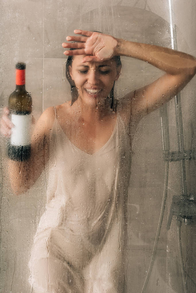 ワインボトルを持ち、ガラスのドアに触れ、シャワーで泣く孤独な落ち込んだ女性  - 写真・画像