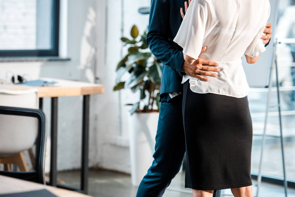 обрізаний вид бізнесмена, що обіймає бізнес-леді, стоячи і фліртуючи в офісі
 - Фото, зображення