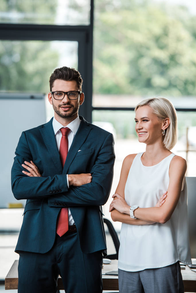 χαρούμενος επιχειρηματίας σε γυαλιά και ελκυστική επιχειρηματίας στέκεται με σταυρωτά μπράτσα  - Φωτογραφία, εικόνα