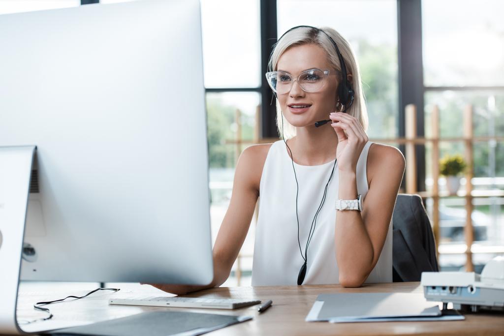 selectieve focus van vrolijke blonde vrouw in de headset werken in Office in de buurt van computer monitor  - Foto, afbeelding
