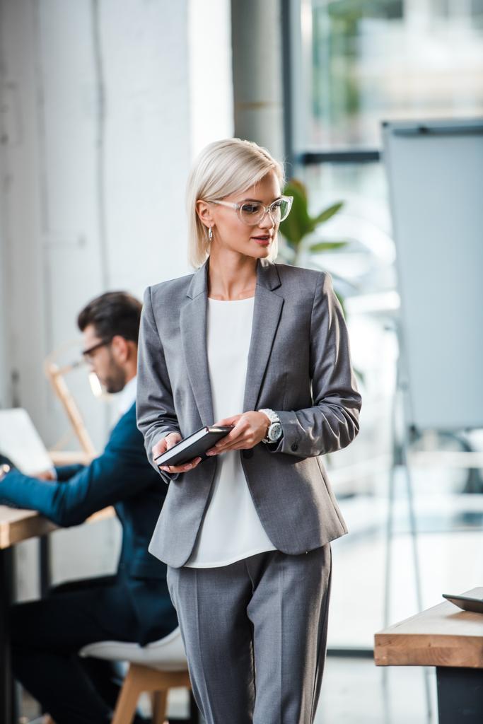 enfoque selectivo de mujer de negocios rubia en gafas con cuaderno cerca de compañero de trabajo en la oficina
  - Foto, imagen