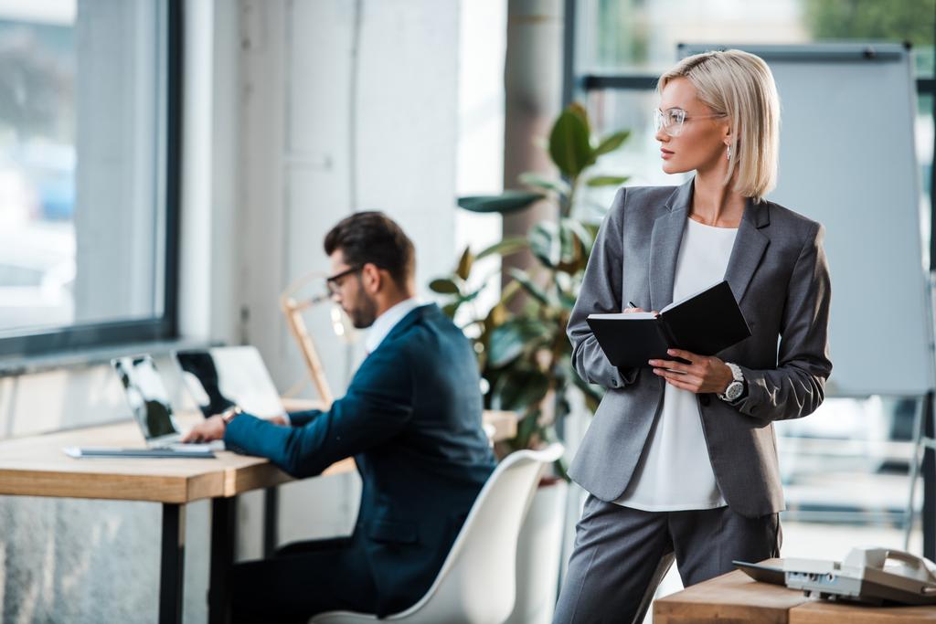 enfoque selectivo de mujer de negocios rubia sosteniendo cuaderno cerca de compañero de trabajo en la oficina
  - Foto, Imagen