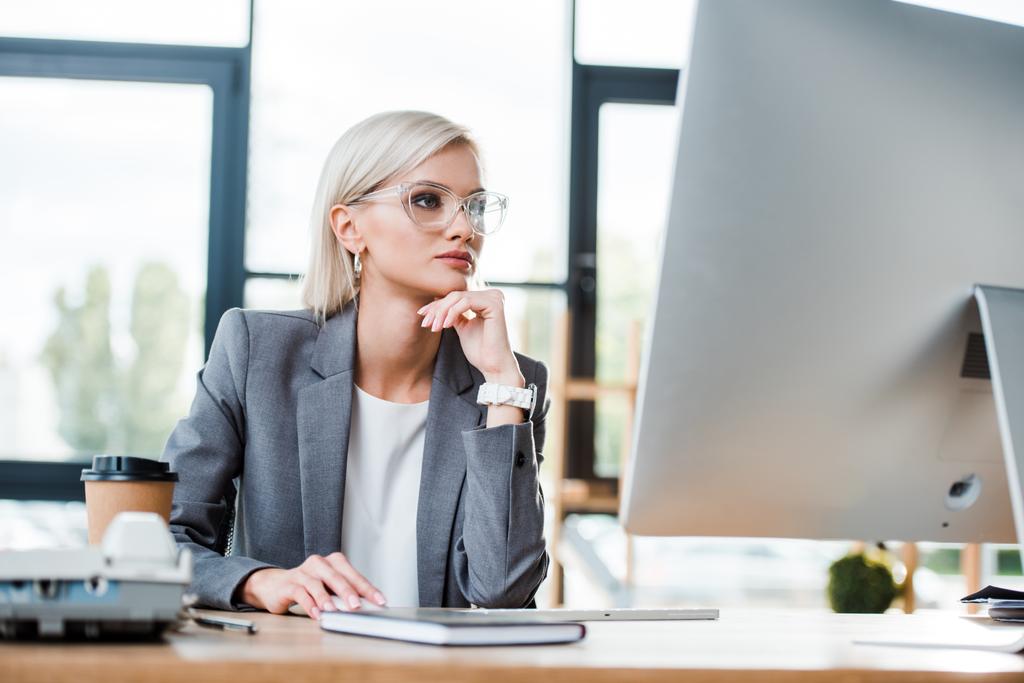 aantrekkelijke blonde vrouw in glazen kijken naar computer monitor in Office  - Foto, afbeelding