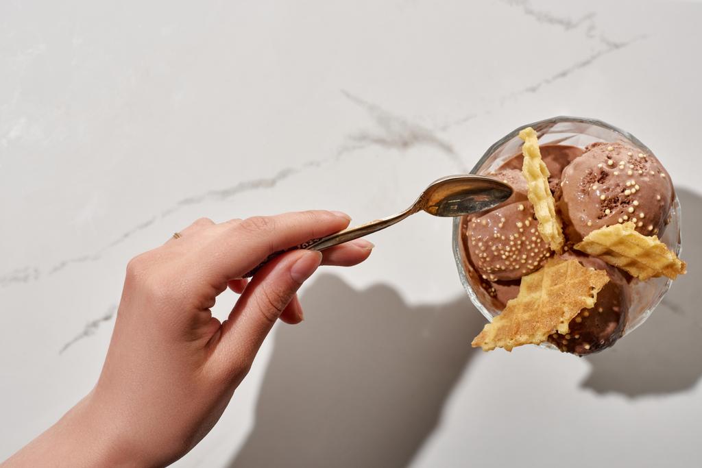cropped view of woman holding soon near delicious chocolate ice cream in bowl with waffles on marble background - Photo, Image