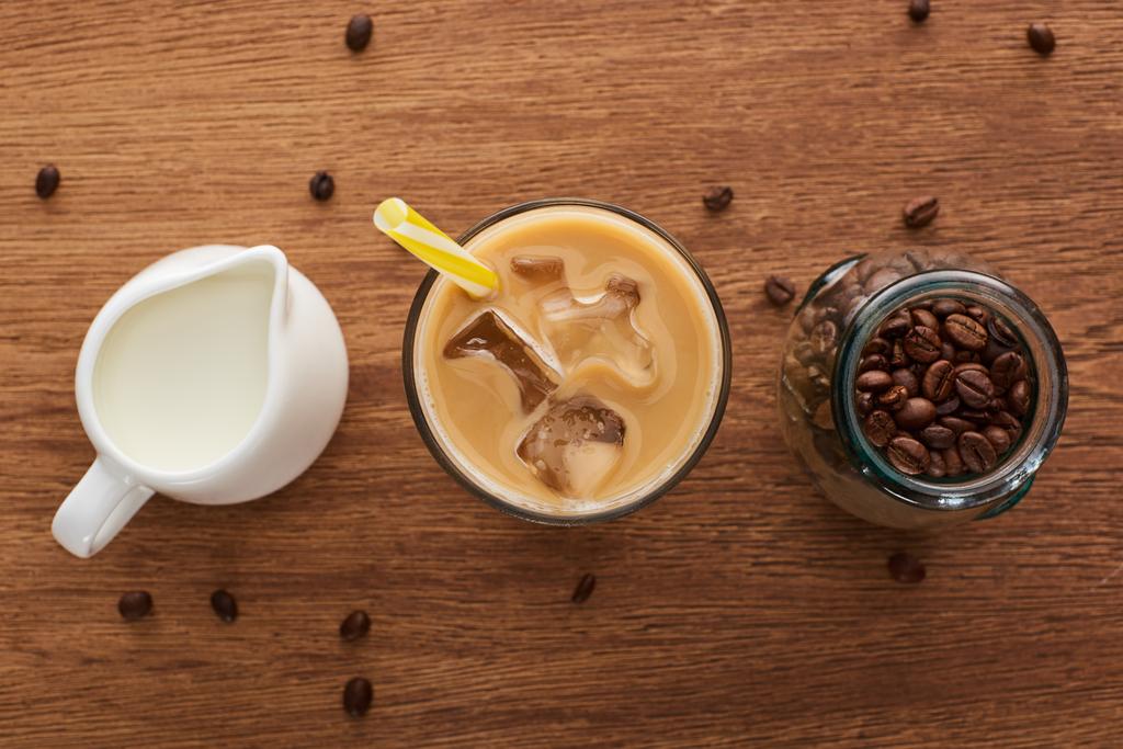 木製のテーブルの上の瓶に水差しとコーヒー粒のミルクの近くにわらとアイスコーヒーのトップビュー - 写真・画像