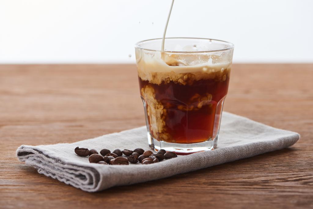 milk pouring into ice coffee in glass on napkin with coffee grains on wooden table isolated on white - Photo, Image